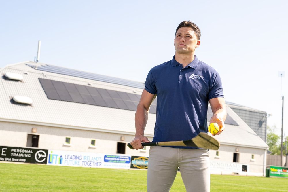 Athlete in navy polo holds tennis racket and ball on grass field with solar-paneled building in background.