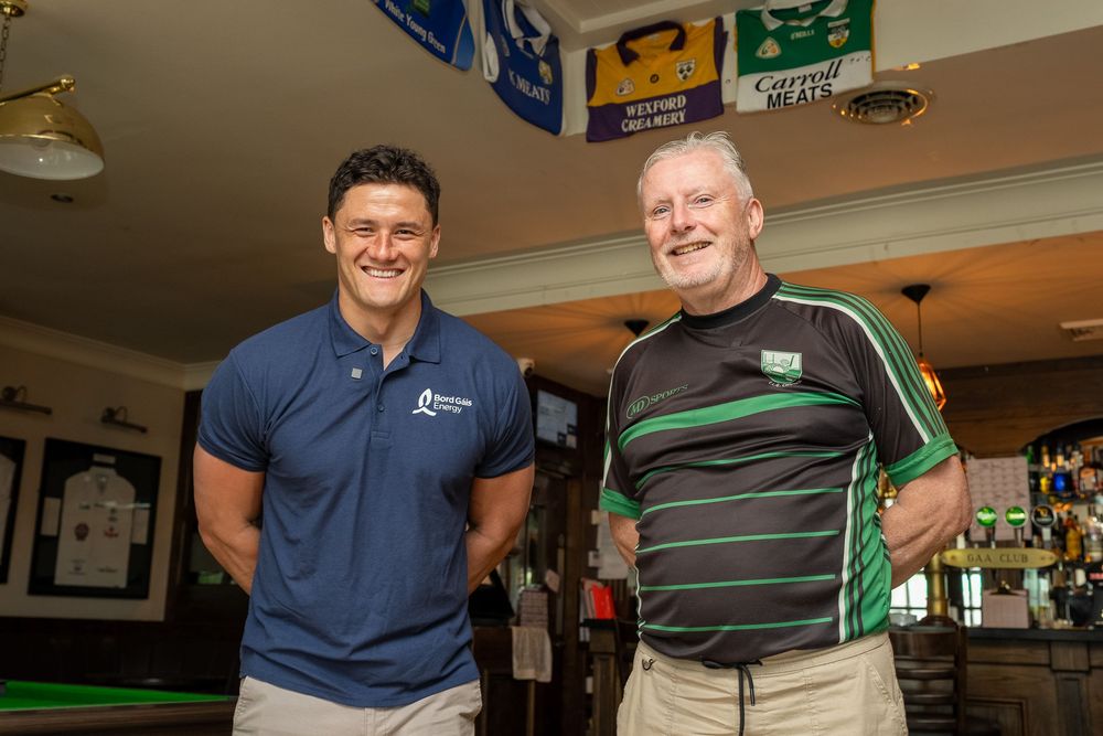 Two men smiling together inside a pub, one in a blue polo shirt and one in a green rugby jersey.