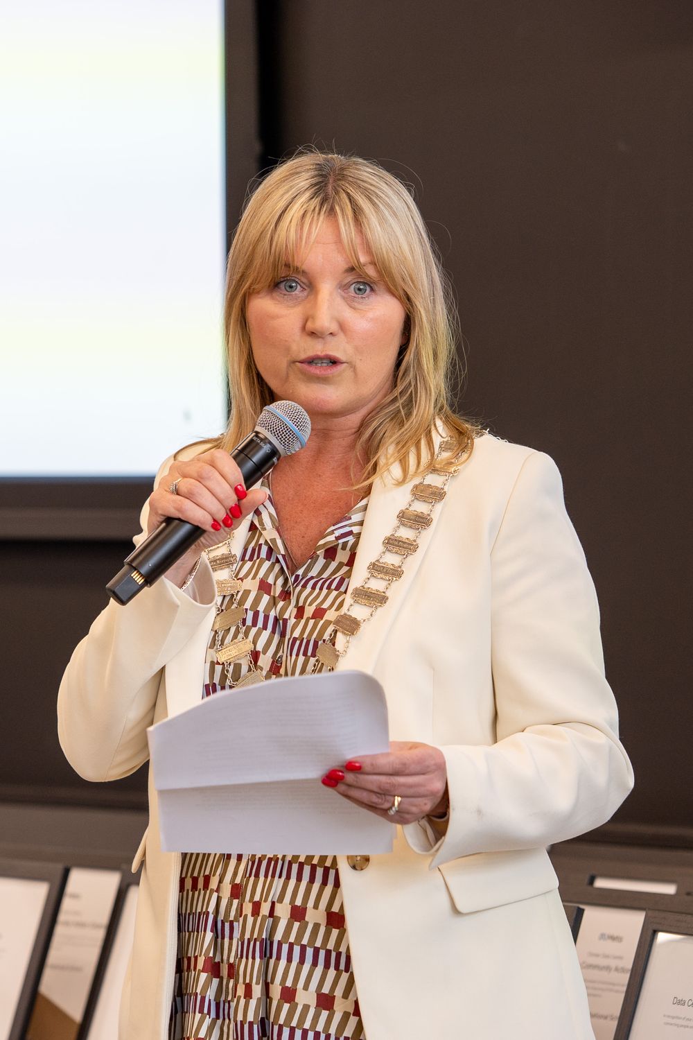 Speaker in white blazer and patterned dress reads from papers at a podium during a presentation.