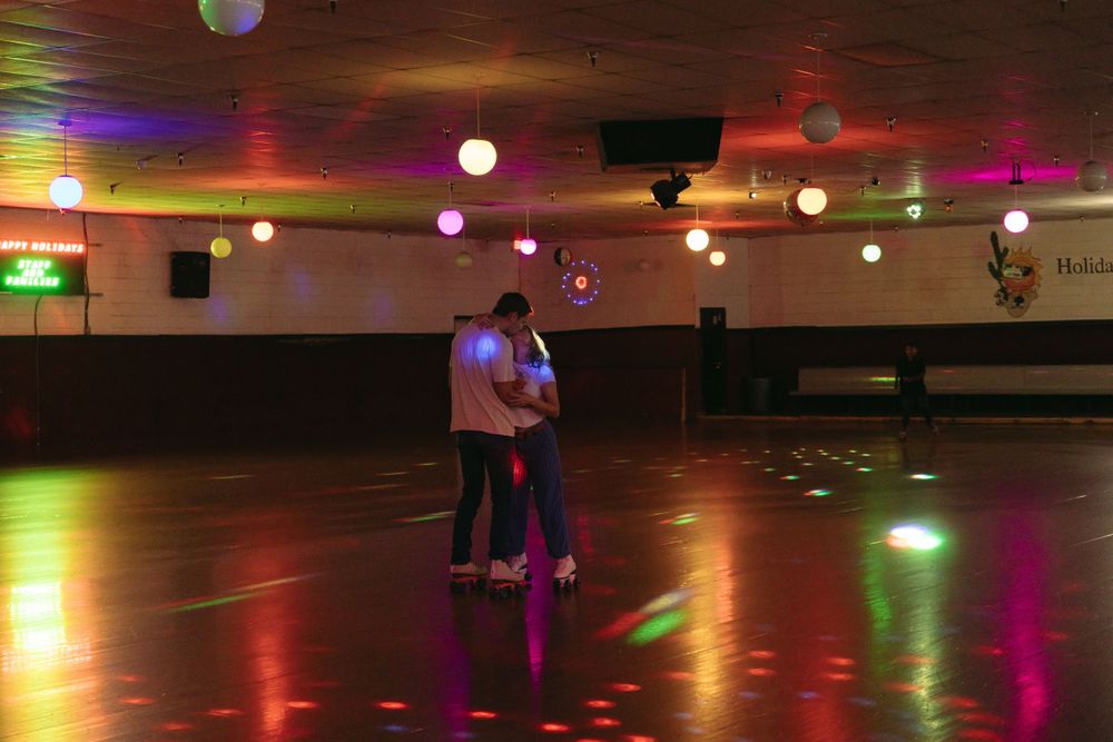 Two people dance together on an empty roller rink floor illuminated by colorful lights and disco balls.