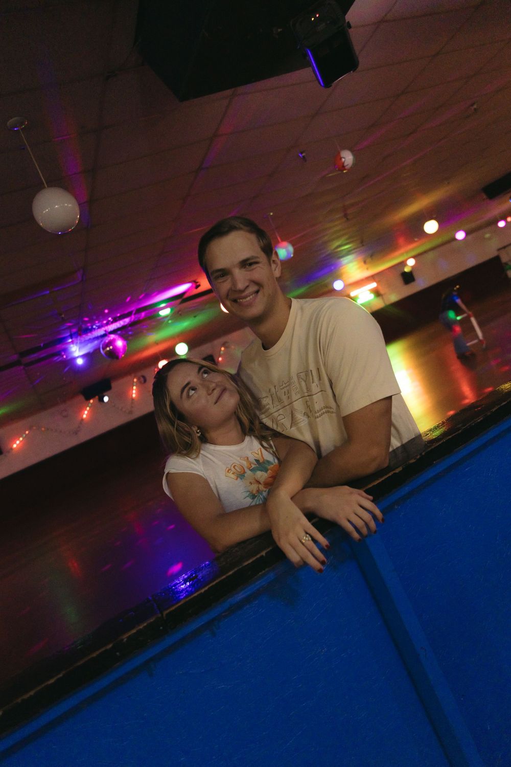 A couple smiles together while leaning on a blue pool table in a colorfully lit entertainment venue.