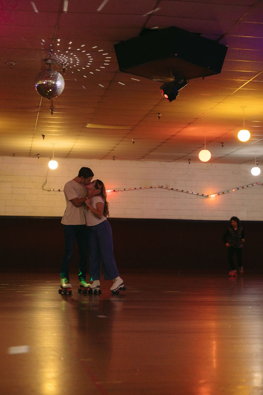 Couple slow dancing on roller skates under a disco ball in a dimly lit roller rink with string lights on the ceiling.