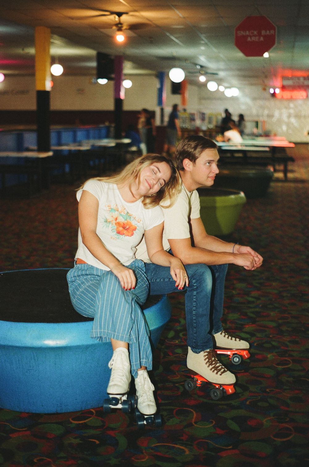 Two people sit together on blue seating at a retro roller skating rink with colorful carpet and vintage lighting.