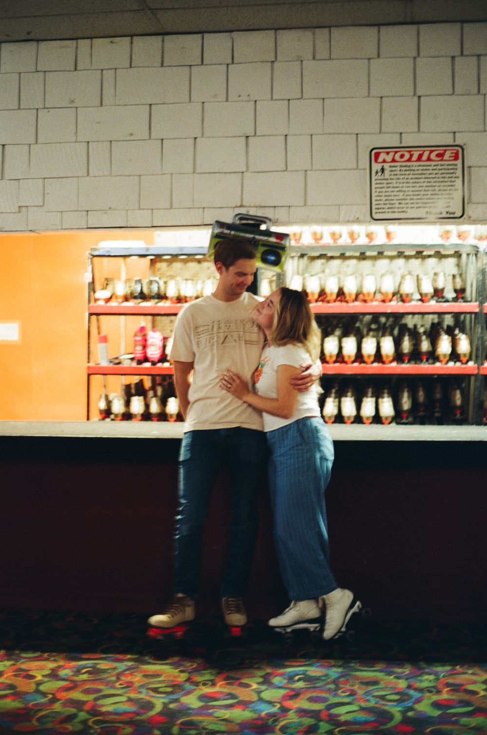 A couple embraces at vintage roller rink with rows of skates and colorful carpeting in the background.