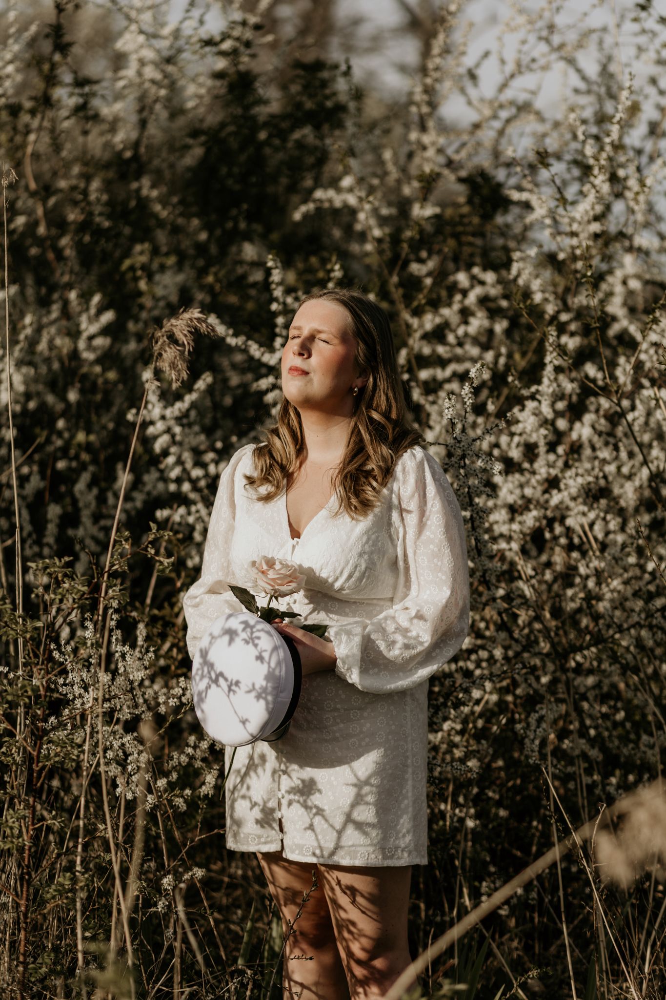 A person in a white long-sleeve dress stands among blooming spring bushes during golden hour.