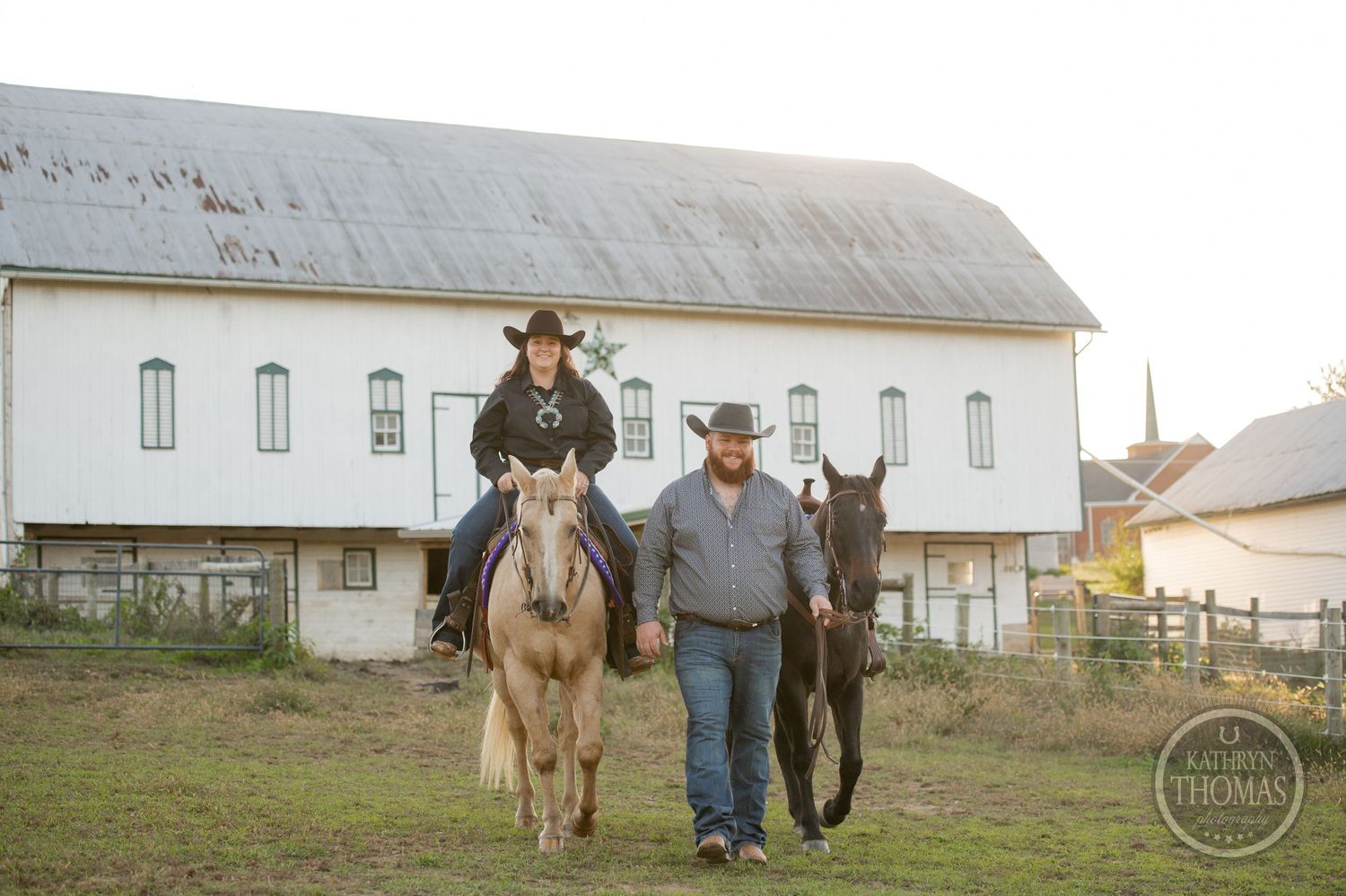 Two horses and people walk together in front of a rustic white barn building.
