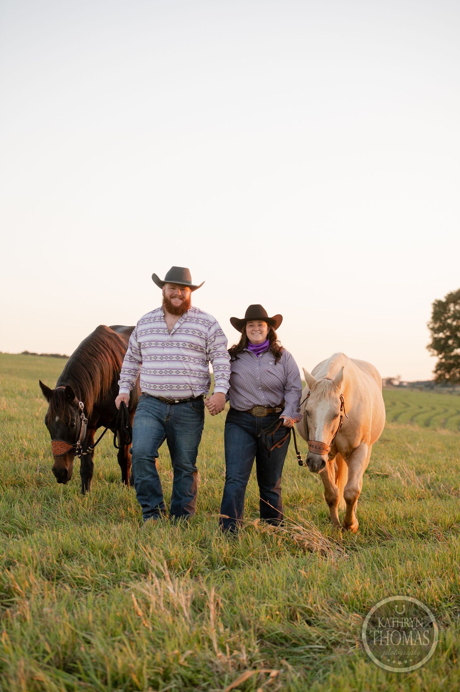 A couple in cowboy hats holding hands while walking with their horses in a sunset pasture.