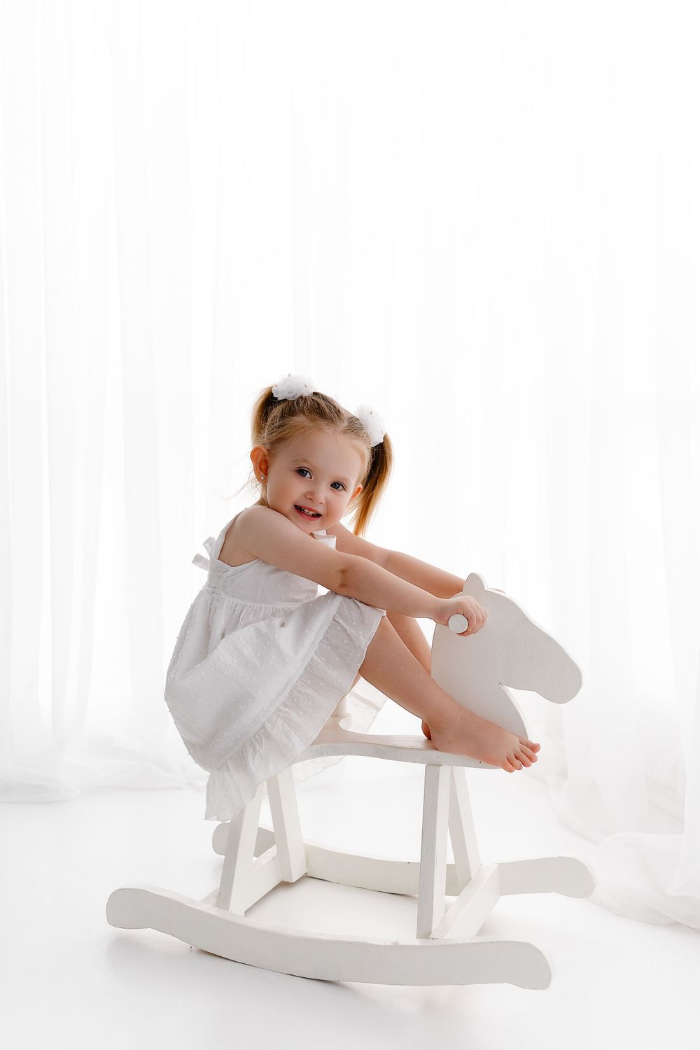 Small child in white dress sitting on decorative white wooden rocking horse against bright background.