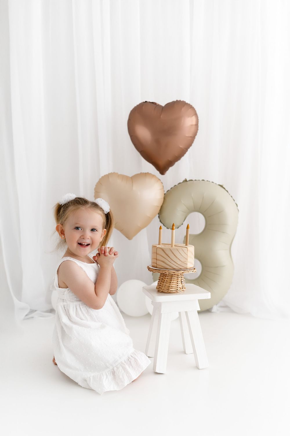 White birthday setup with heart-shaped balloons in rose gold and beige tones behind a small white pedestal with cake.