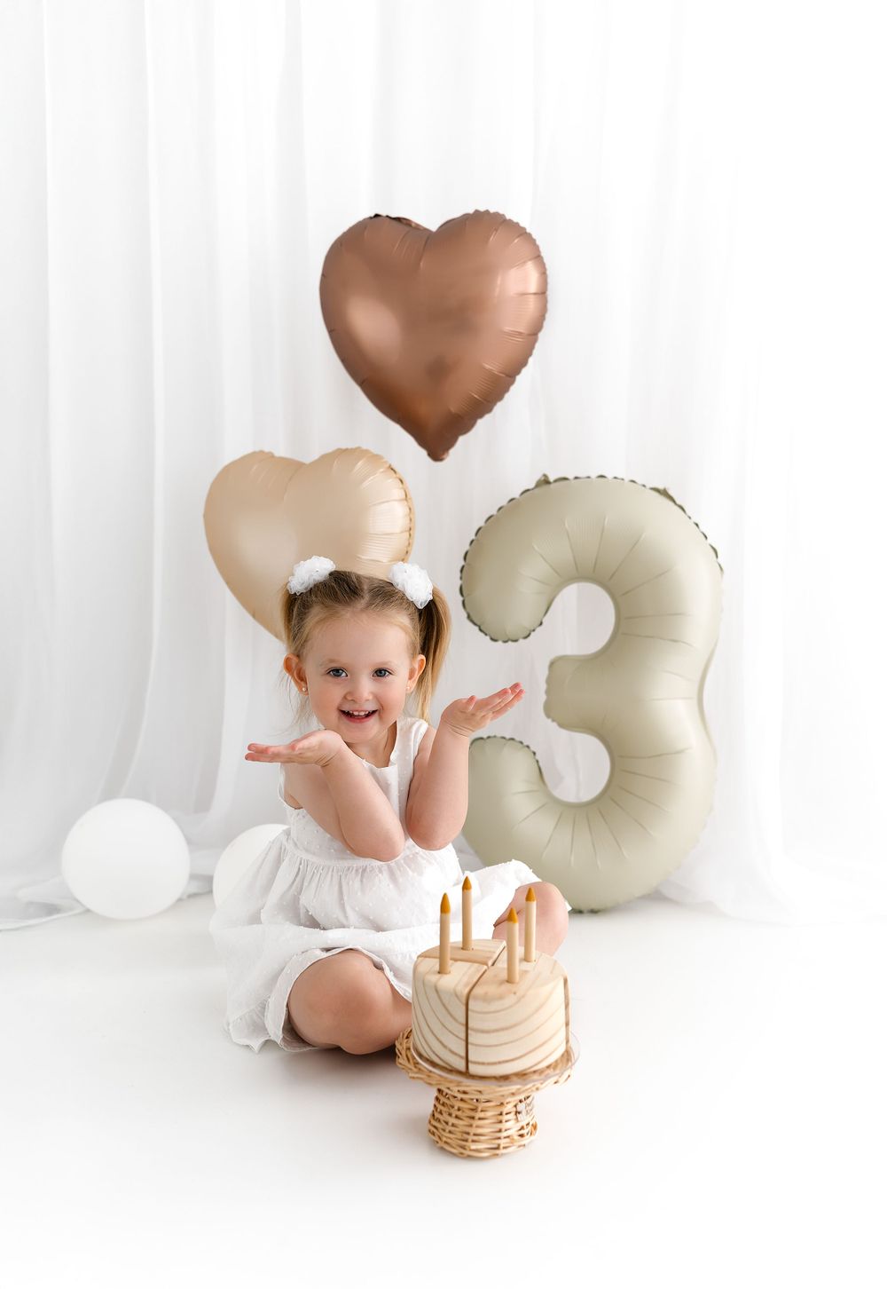 Festive birthday setup with rose gold and cream balloons featuring number 3 and heart shapes beside a wooden cake display.