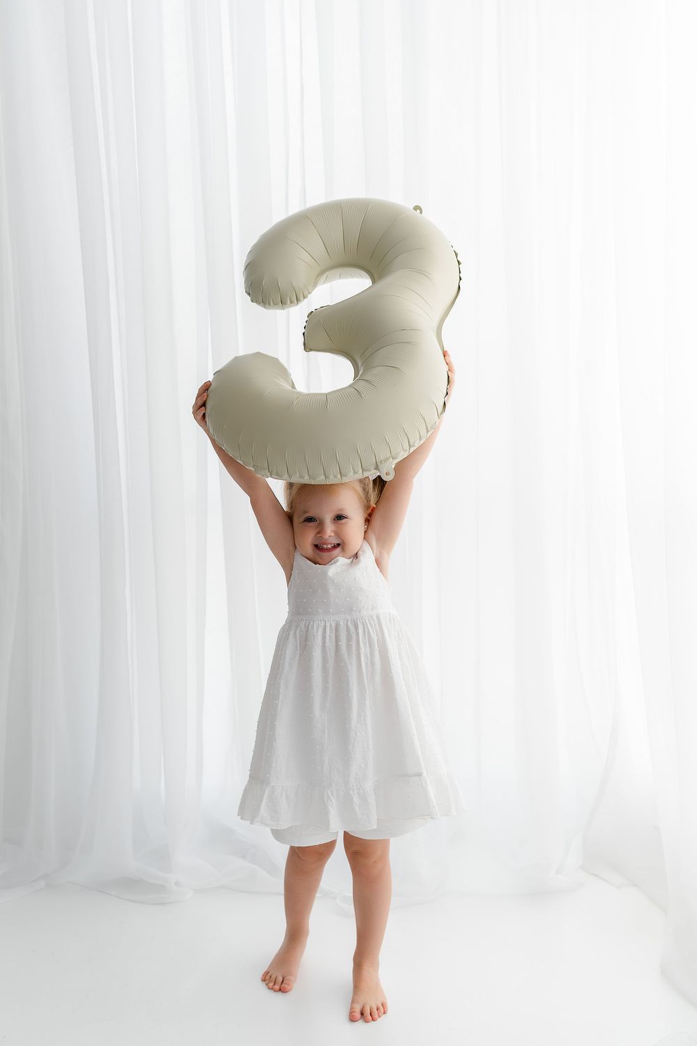Child in white dress holds large inflatable number 3 balloon against white background.