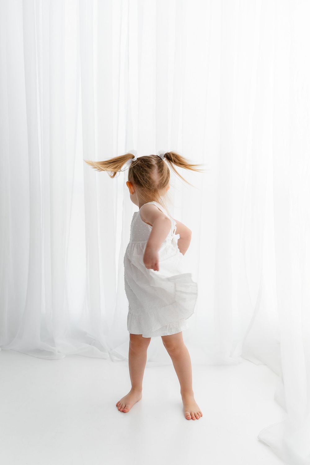 Young child in white dress spins playfully with pigtails flying, standing barefoot against white curtain backdrop.