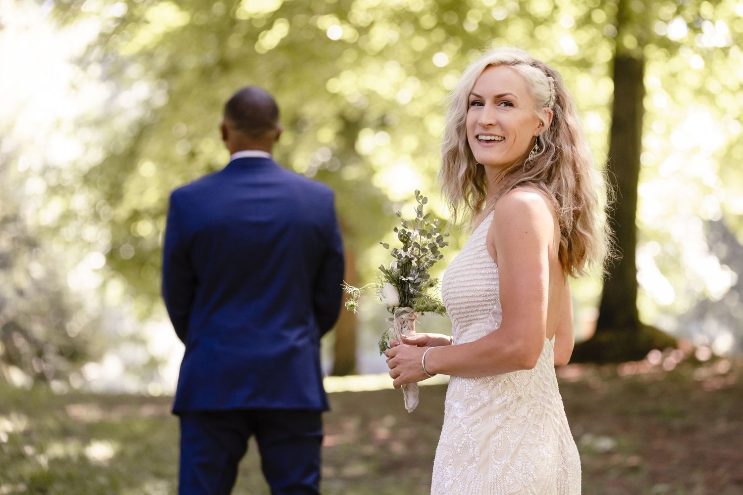 Bride holding a bouquet laughs joyfully during first look photos in a sunlit forest setting.