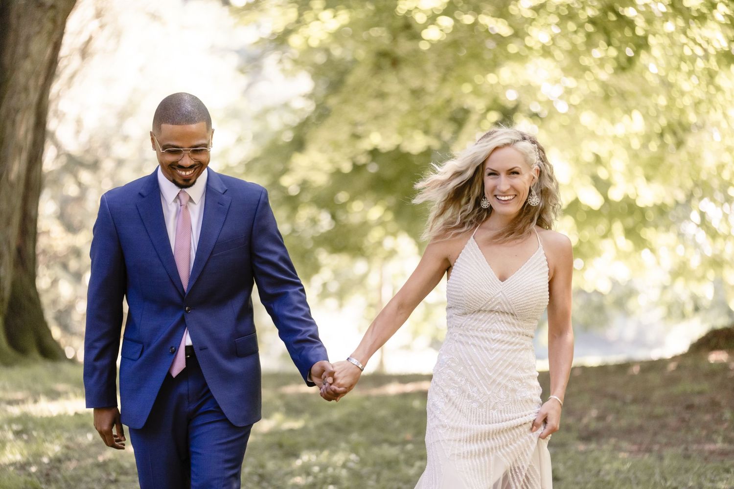 Joyful newlyweds hold hands while walking through a sun-dappled garden setting.