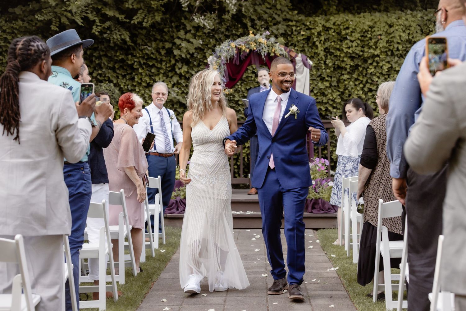 Newlyweds walk back down the aisle as wedding guests celebrate at an outdoor garden ceremony.