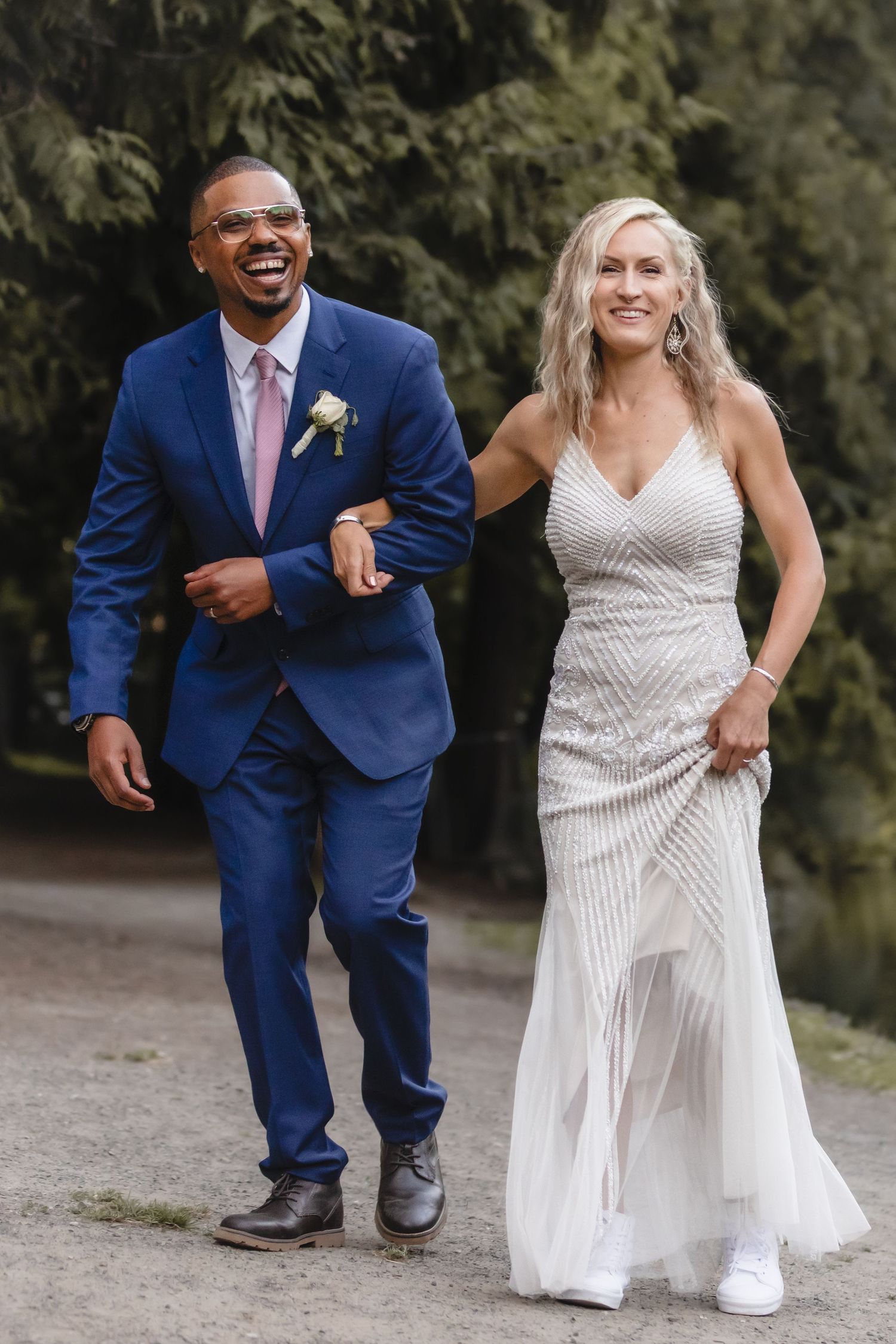 A happy couple shows off their wedding attire while walking together on a scenic outdoor path.