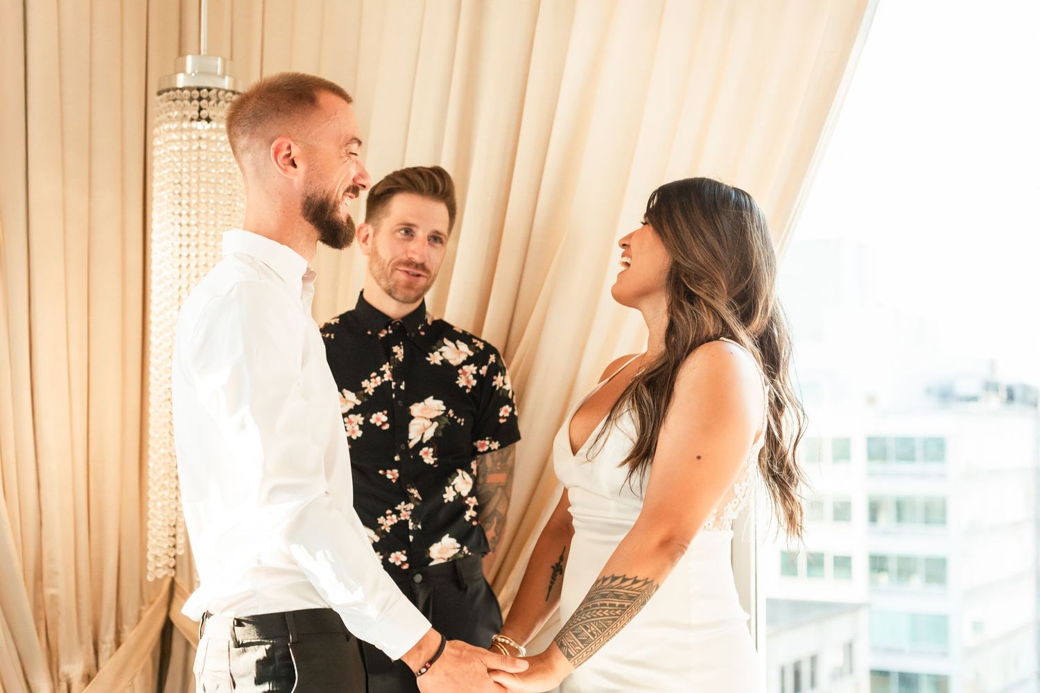 A wedding ceremony takes place by a window with beige curtains and city views in the background.