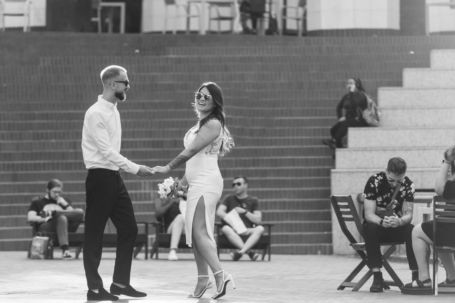 A couple dances together on a plaza with onlookers sitting on steps in the background in black and white photography.