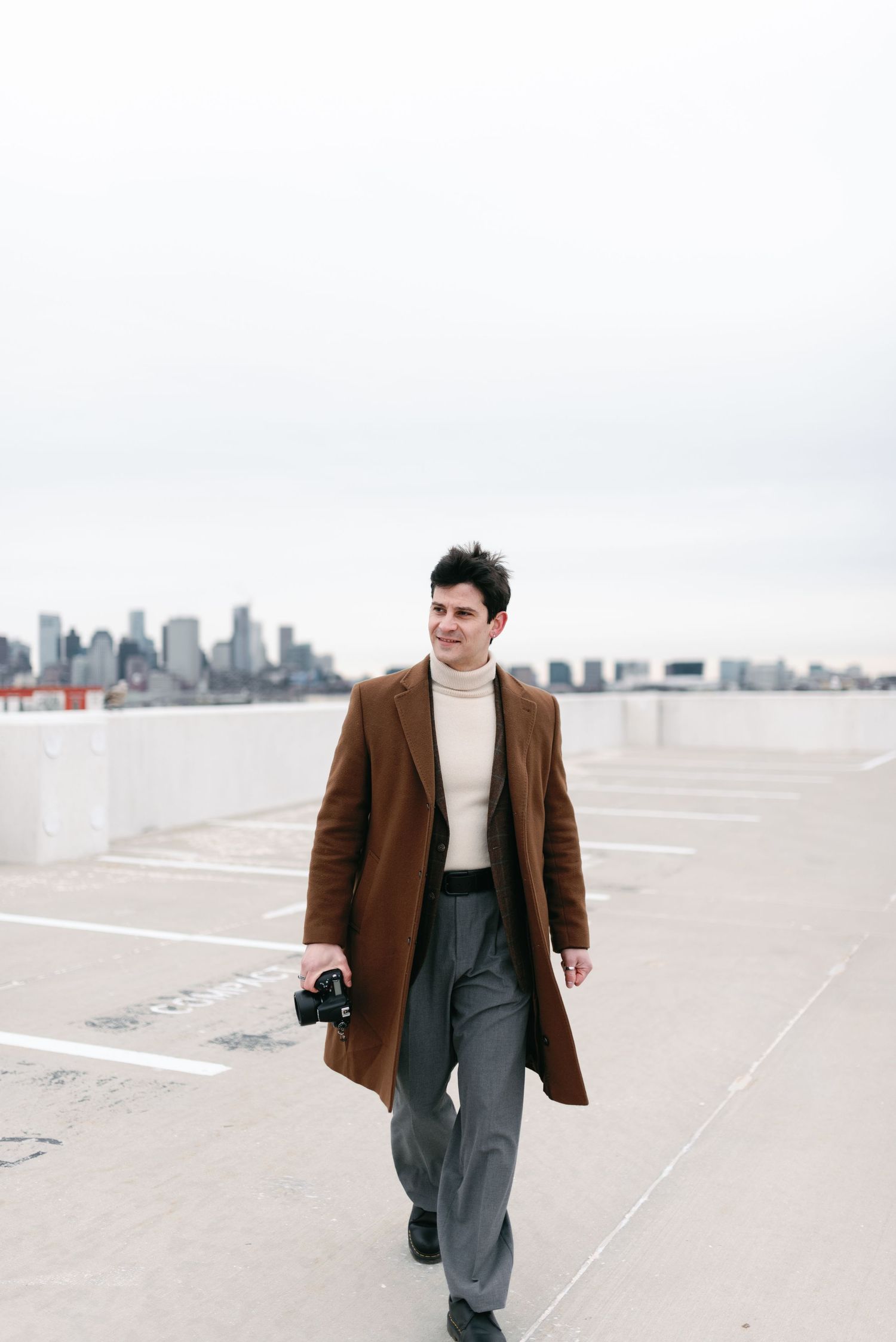 A photographer in a brown coat walks along a city rooftop holding a camera.