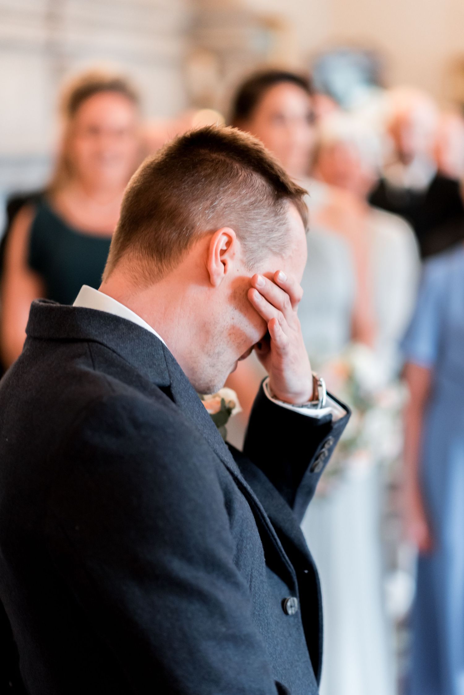 Emotional groom in dark suit covers his face during wedding ceremony while guests watch in background.