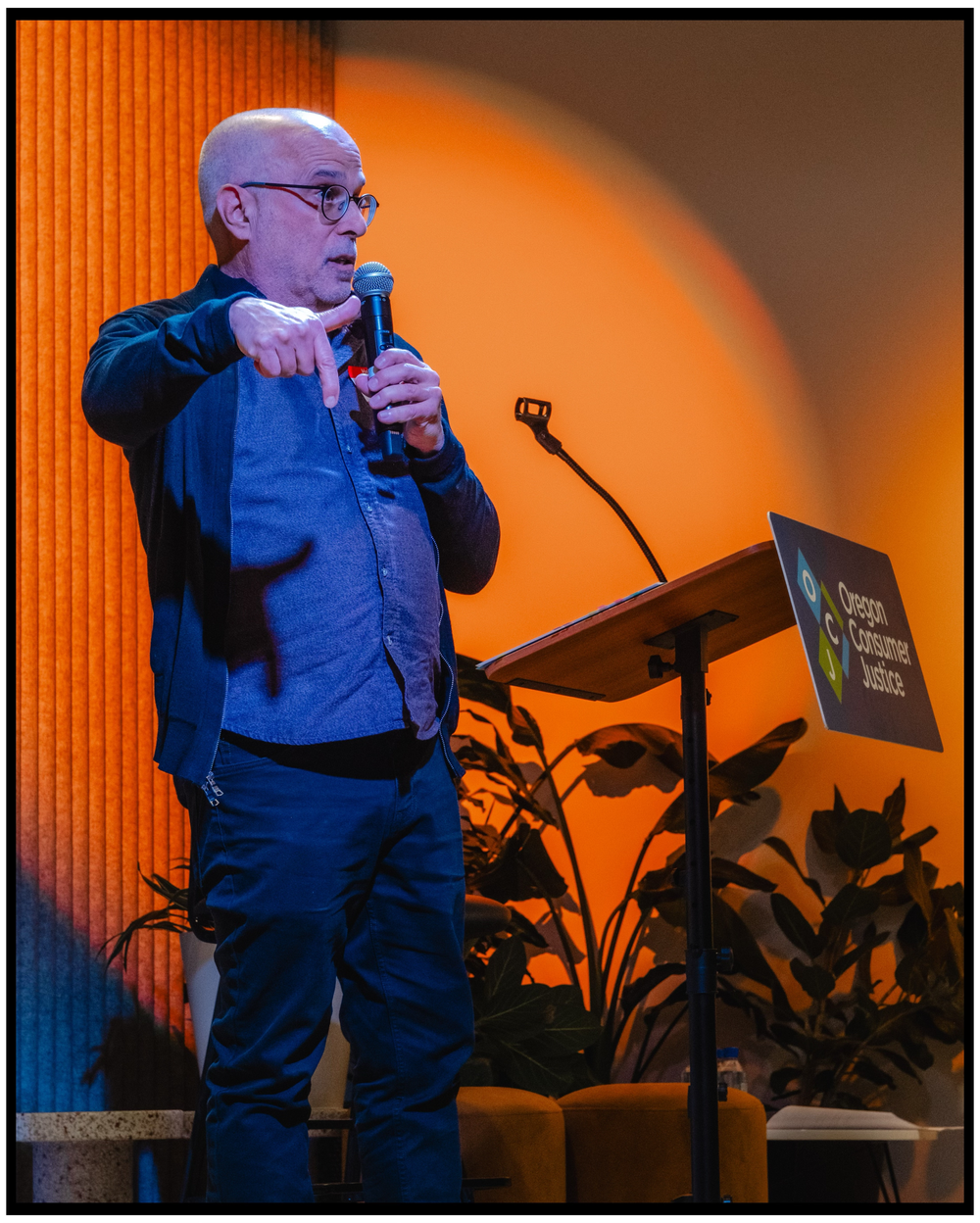 Speaker presenting on stage with orange backdrop and potted plants for ambiance. Portland event photographer
