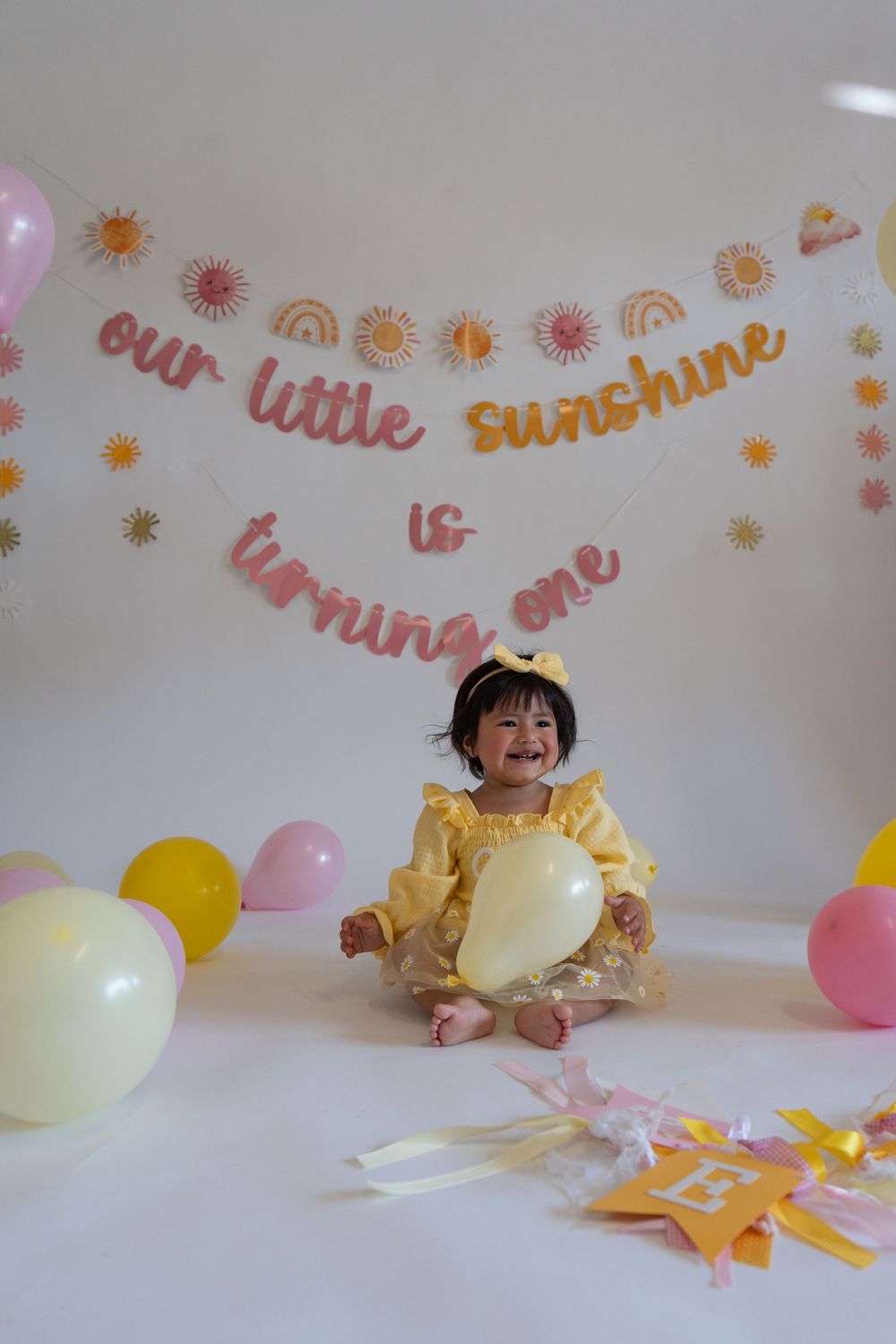 A child plays with colorful balloons and decorations at a party with pink and yellow themed decor and text on the wall.