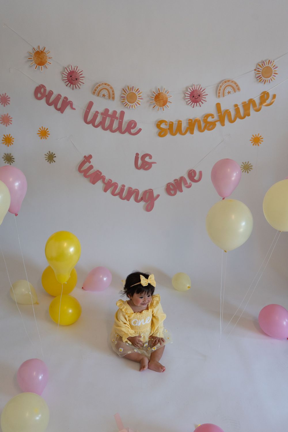 Festive birthday setup with pink and yellow balloons against white wall with sunshine-themed decorations and text.