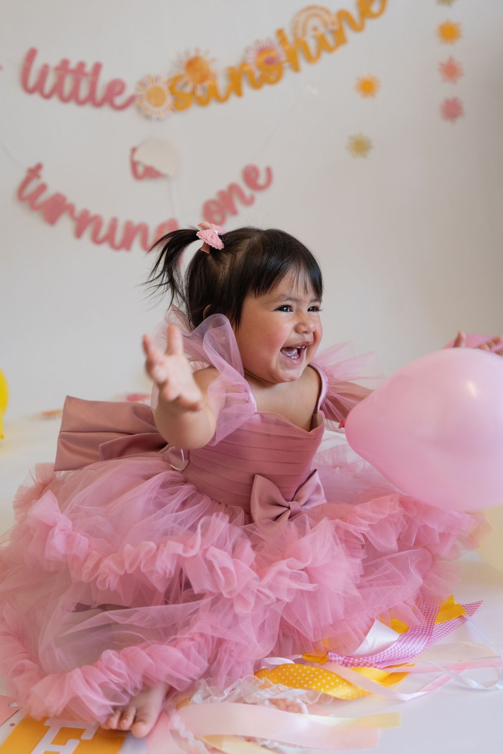 A happy baby wearing a pink tulle dress laughs while lying on a decorated floor with balloons and decorations.