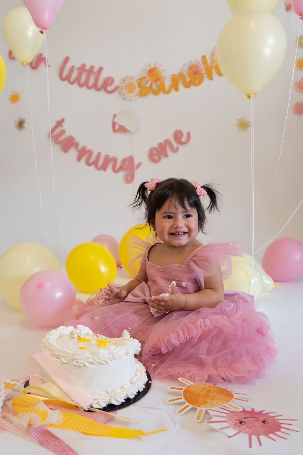 A cake smash photo session with pink tulle dress and yellow and pink balloons for a special celebration.