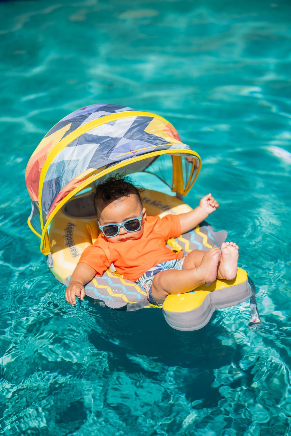 Small child floating in yellow and gray pool float wearing orange vest and sunglasses.