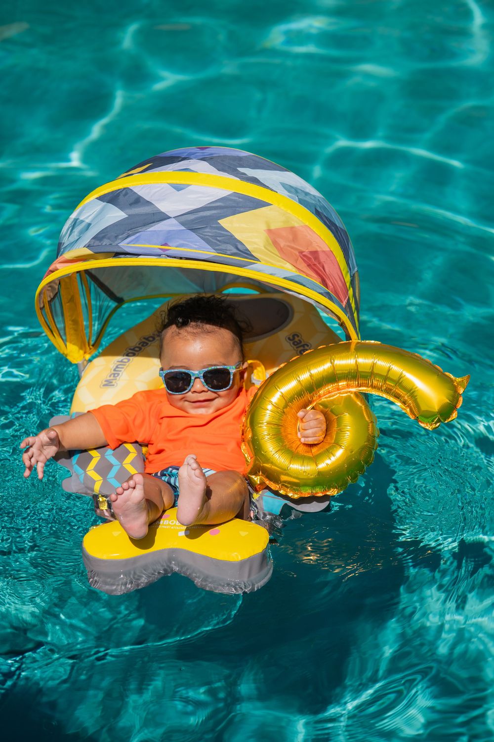 Baby swimming in turquoise pool with gold balloon and colorful float.