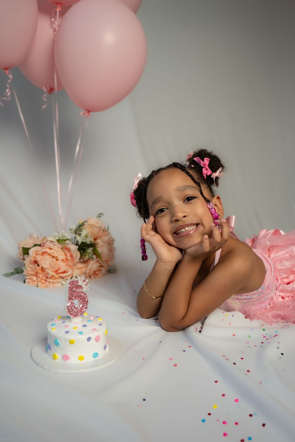 Birthday cake smash photoshoot with pink balloons and confetti on grey background.