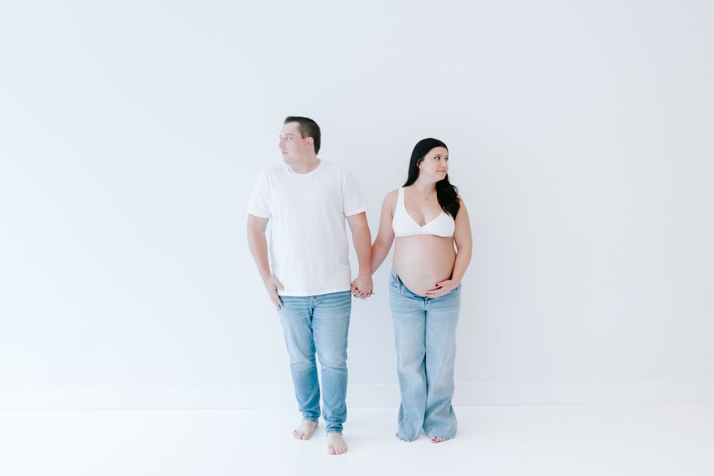 Minimalist maternity portrait with couple in casual wear against white wall.
