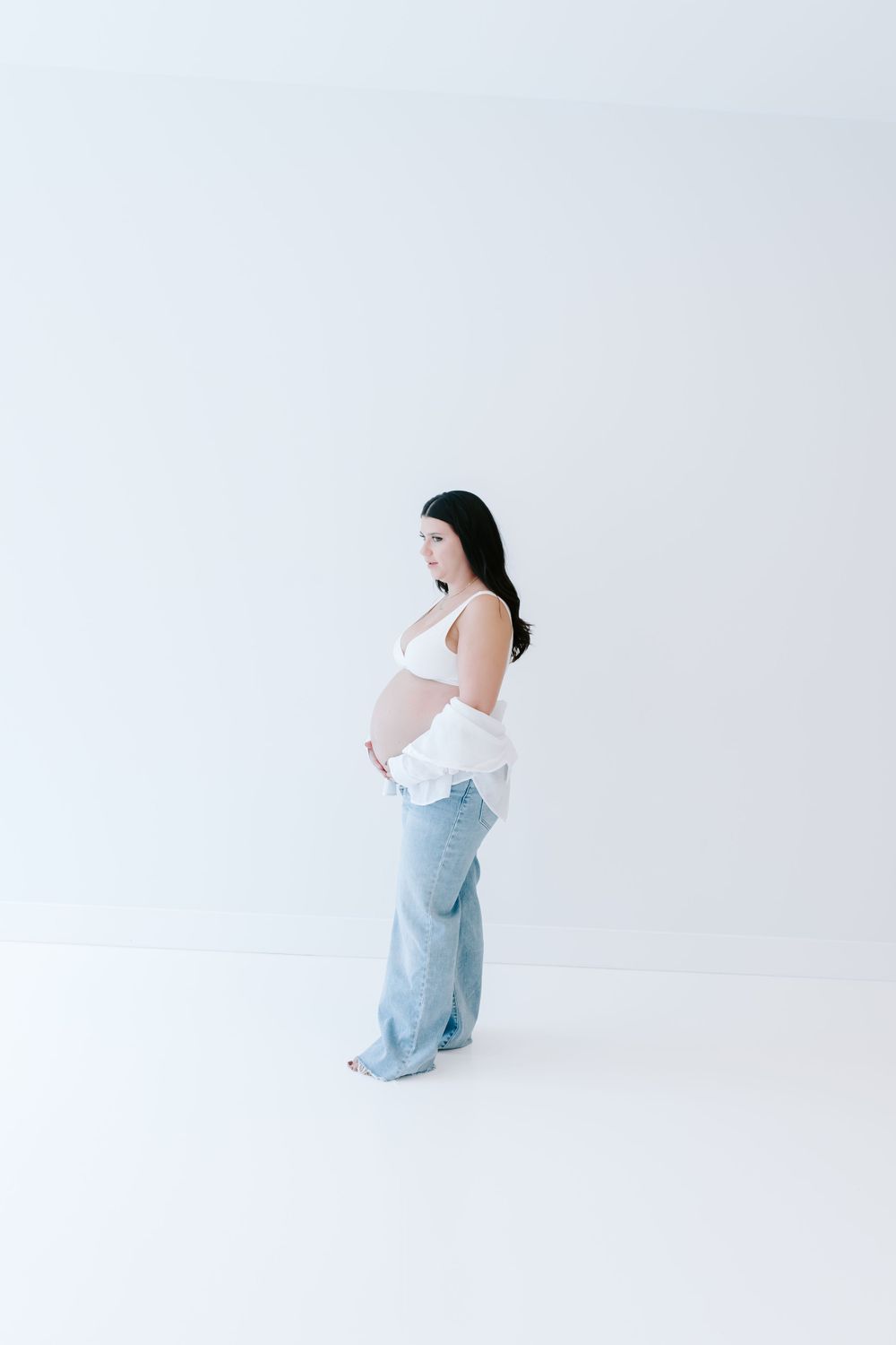 Side view maternity portrait against white backdrop showing expecting mother in casual denim.