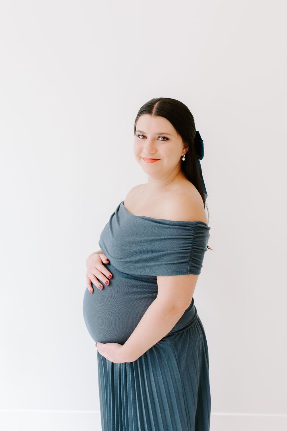 Elegant maternity pose in blue off-shoulder pleated dress against white background.