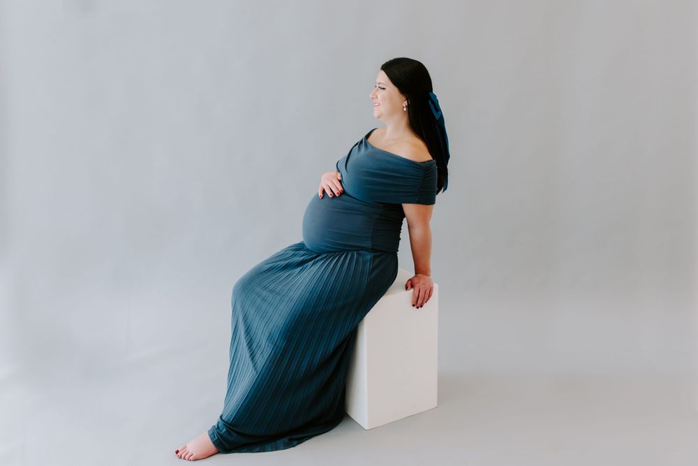 Pregnant woman in blue dress sitting on white cube in studio setting.