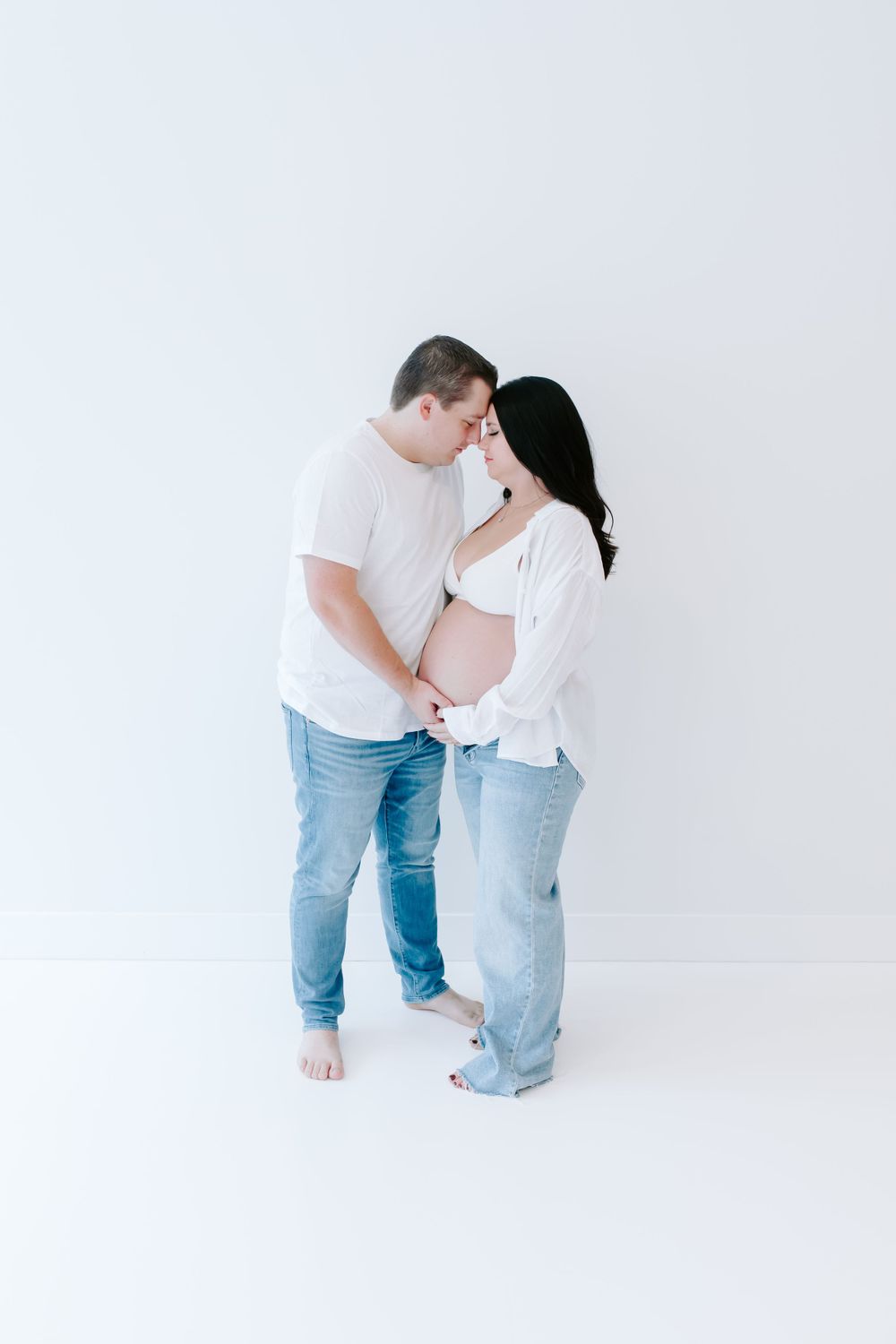 Intimate maternity portrait of couple in white and denim sharing a tender moment against white backdrop.