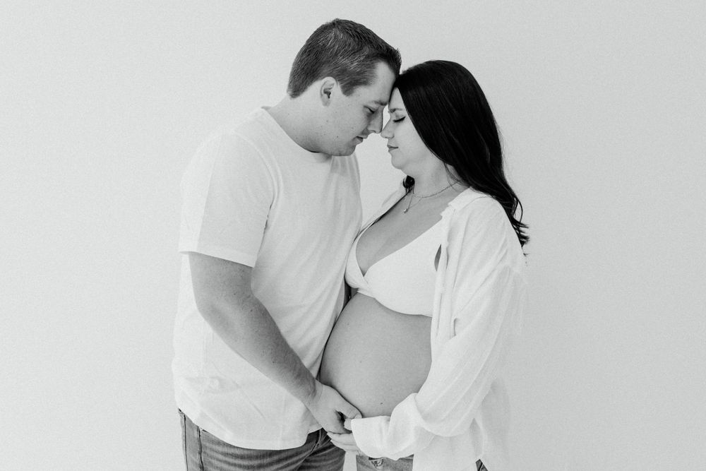 Intimate black and white maternity photo with couple in white against plain backdrop.
