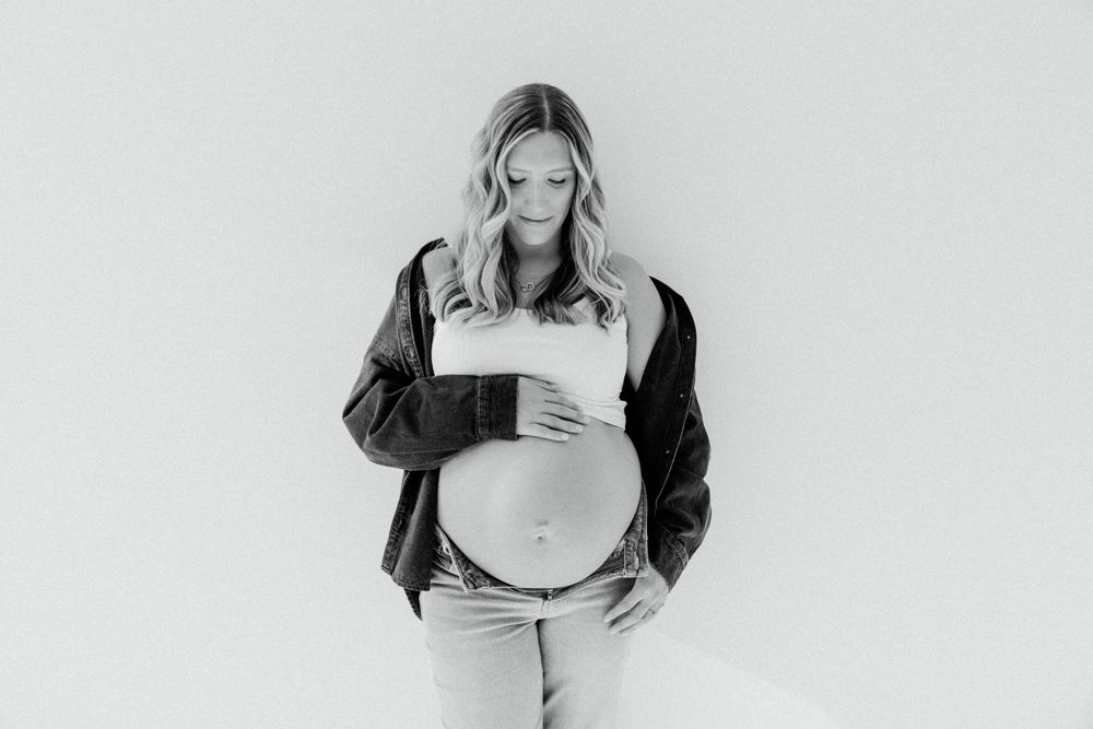 Black and white maternity photo of an expectant mother wearing a leather jacket against a white wall.