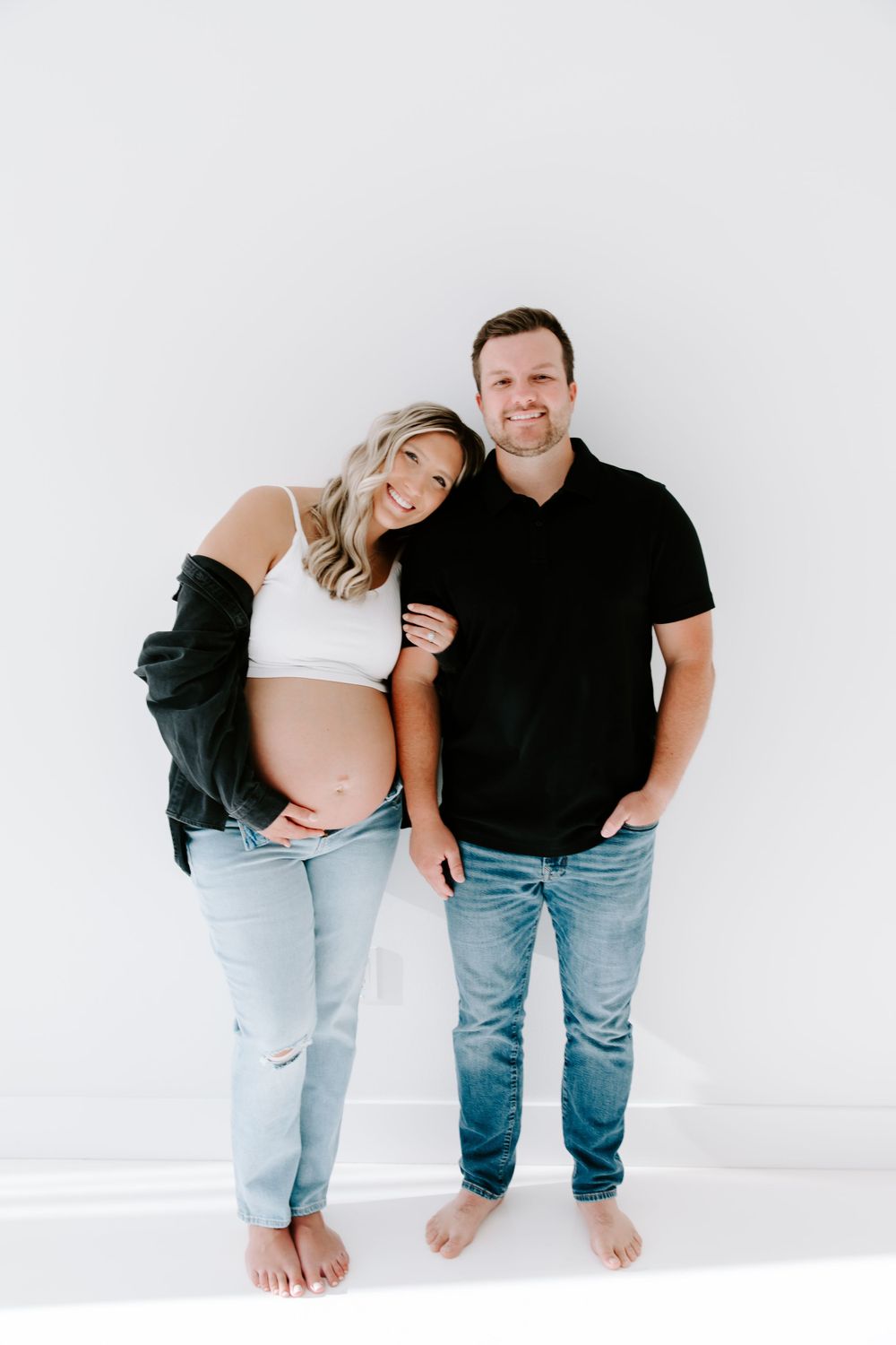 Casual maternity portrait of a couple in denim against a white backdrop.