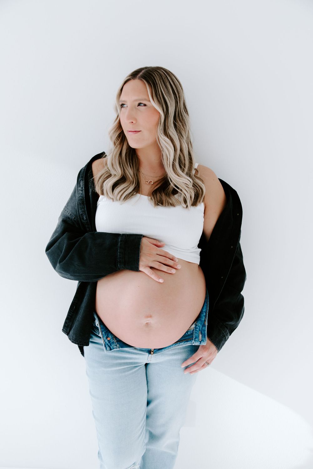 Modern maternity photo with black jacket and white crop top against minimal background.