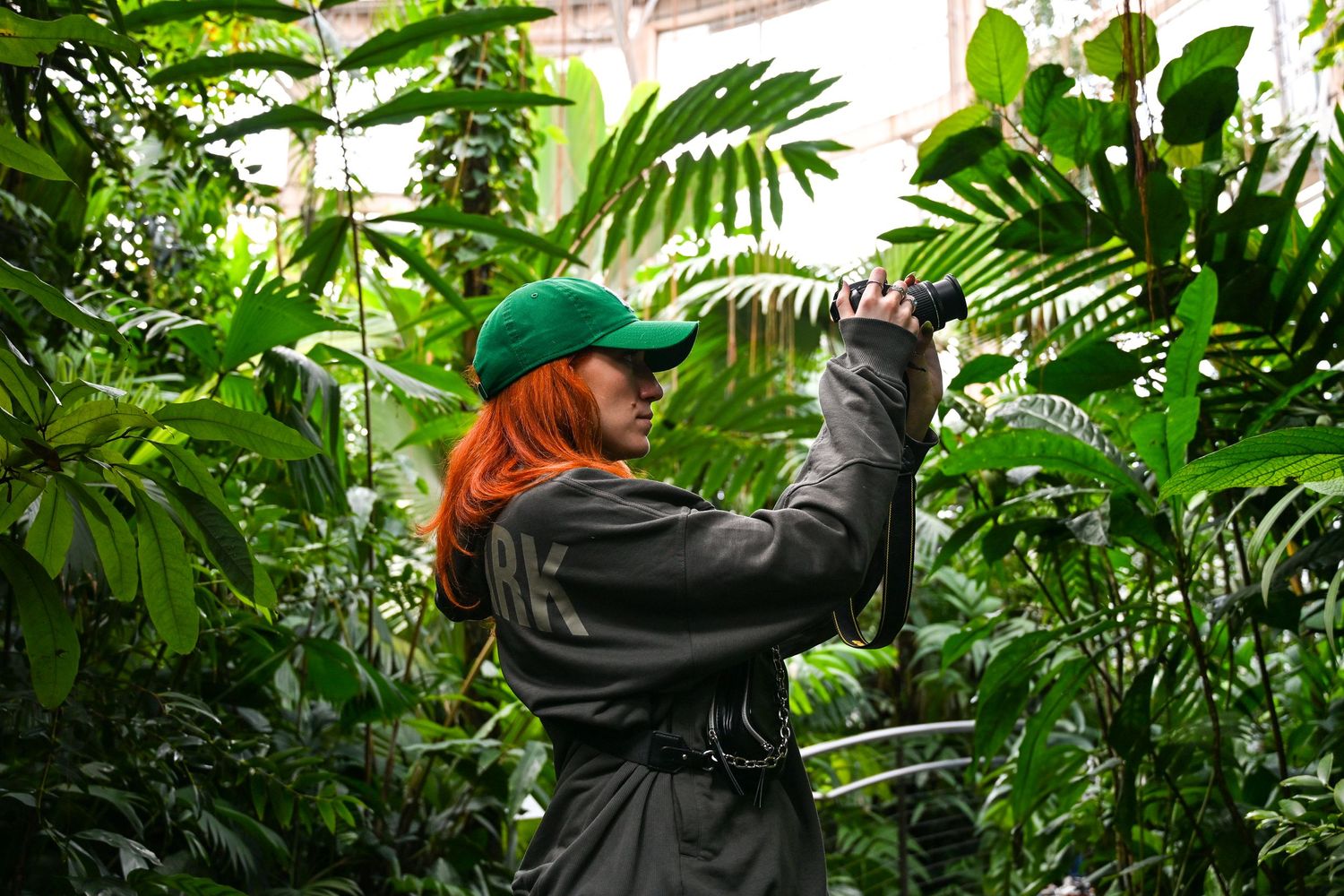 Someone wearing a green cap and gray jacket takes a photo while surrounded by lush tropical foliage in a greenhouse.