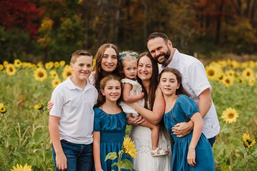 Family of seven smiling together in a sunflower field during autumn, wearing coordinating blue and white outfits.