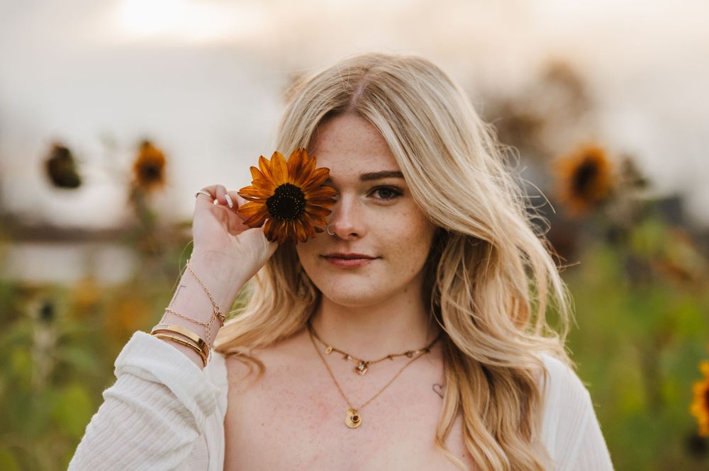 Young blonde woman holding a sunflower over her eye in a sunflower field, wearing a white sweater and gold necklace.