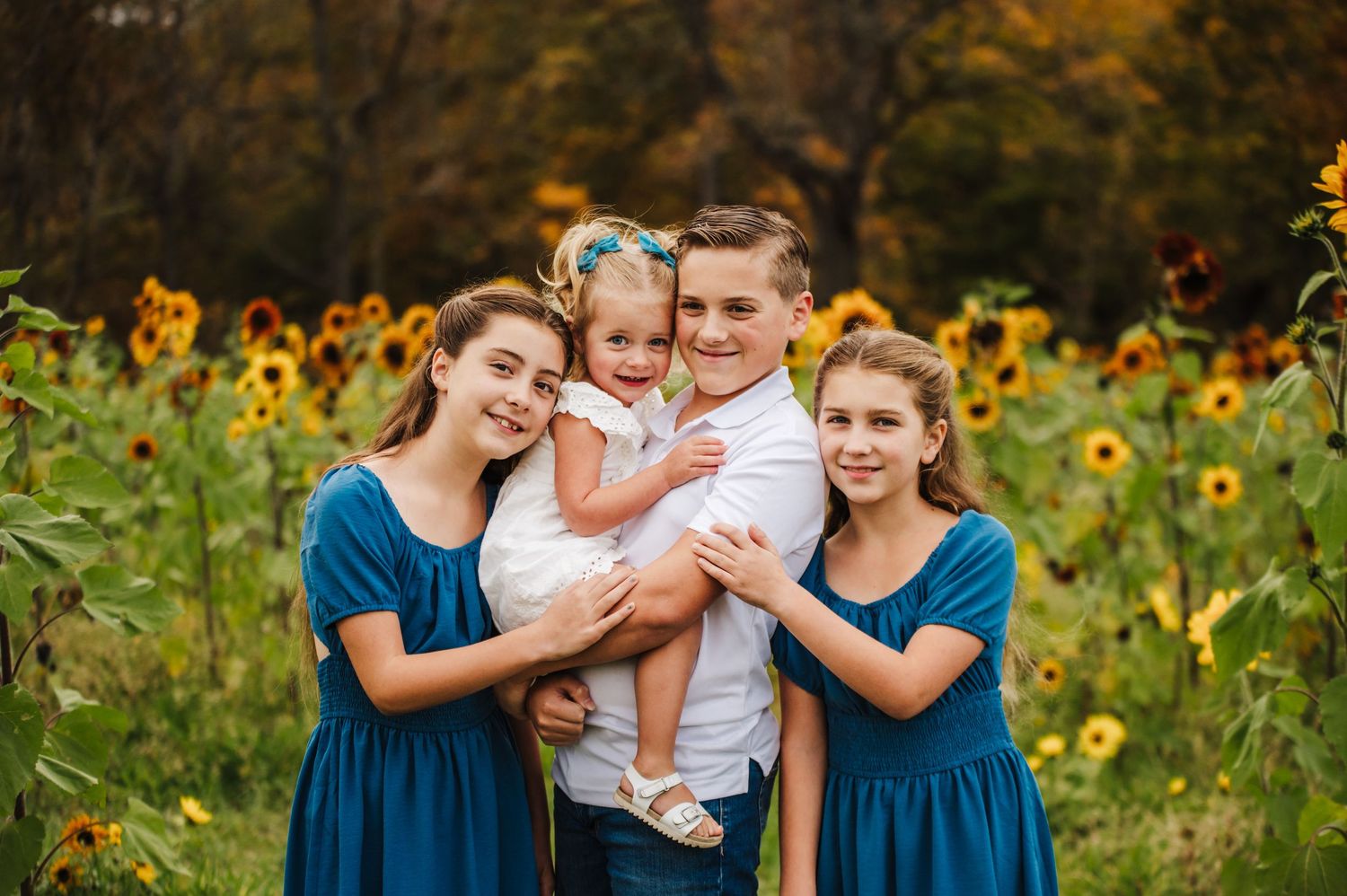 Four siblings smile together in a sunflower field, with the boy holding a toddler girl, flanked by two girls in teal dresses.