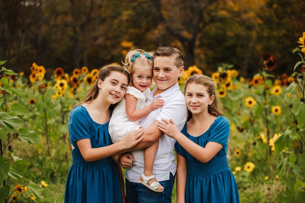 Four siblings smile together in a sunflower field, with the boy holding a toddler girl, flanked by two girls in teal dresses.