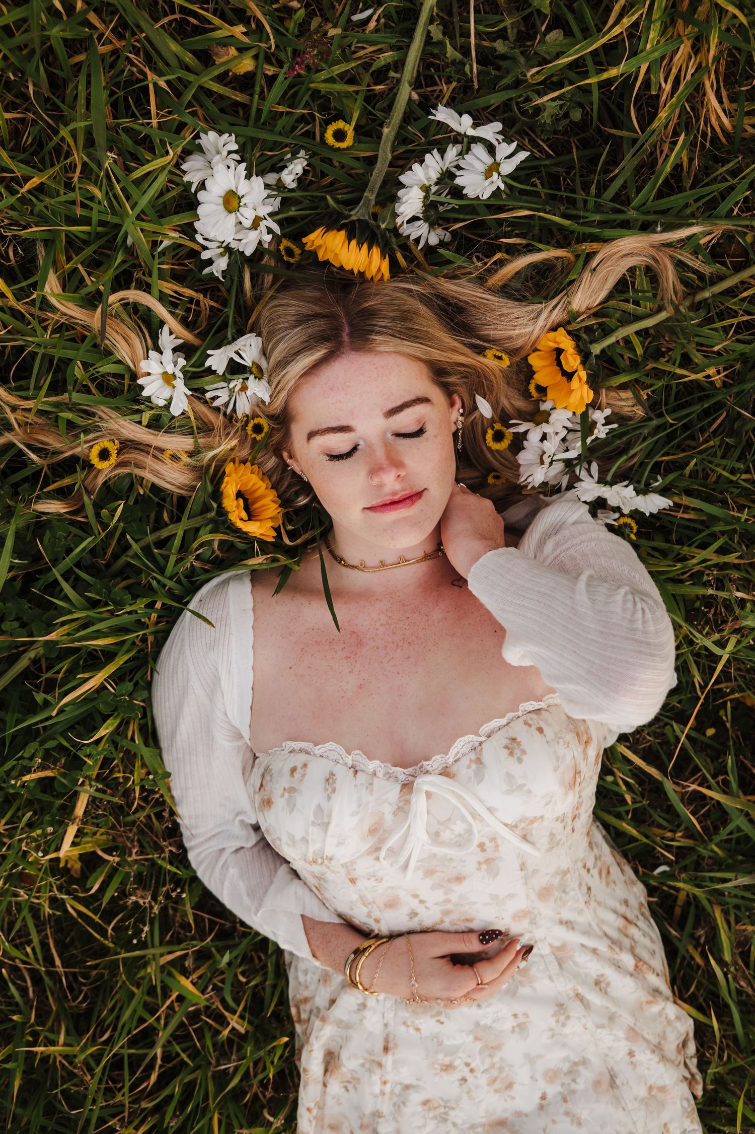 Young woman in floral dress lying in green grass with sunflowers and daisies in her blonde hair.