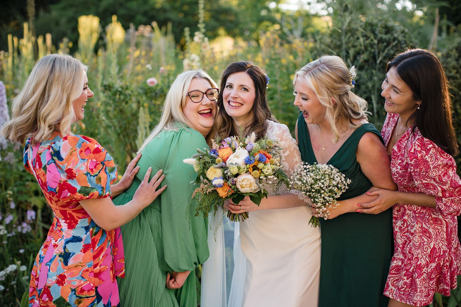A group of women in colorful dresses gather together in a garden setting, laughing and holding a floral bouquet.