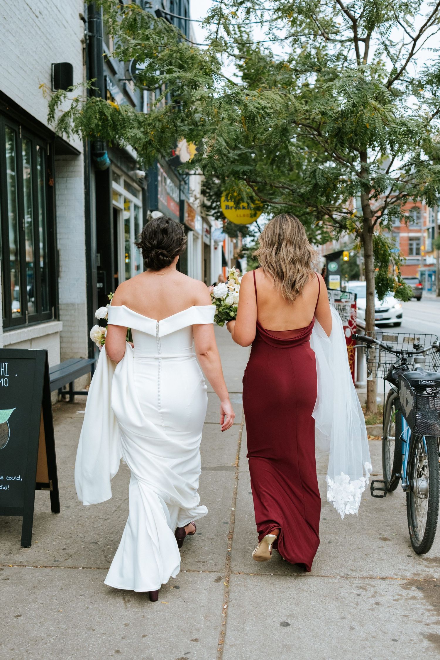 Two people walking down a city sidewalk, one in a white wedding dress and one in a burgundy gown.