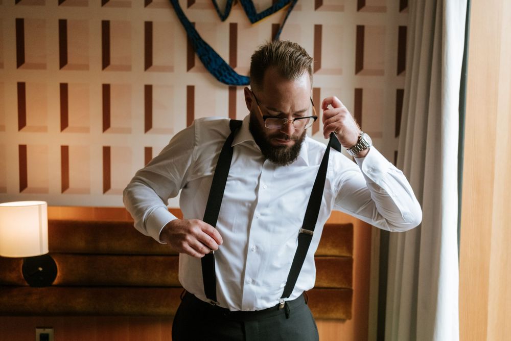 Groom adjusting suspenders while getting ready in white shirt.